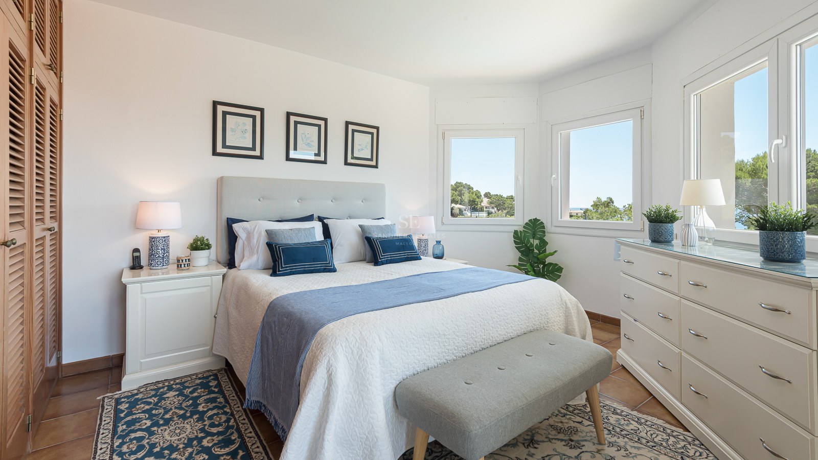 Chambre lumineuse avec lit blanc, coussins bleus, commode blanche, grandes fenêtres et sol en terre cuite.