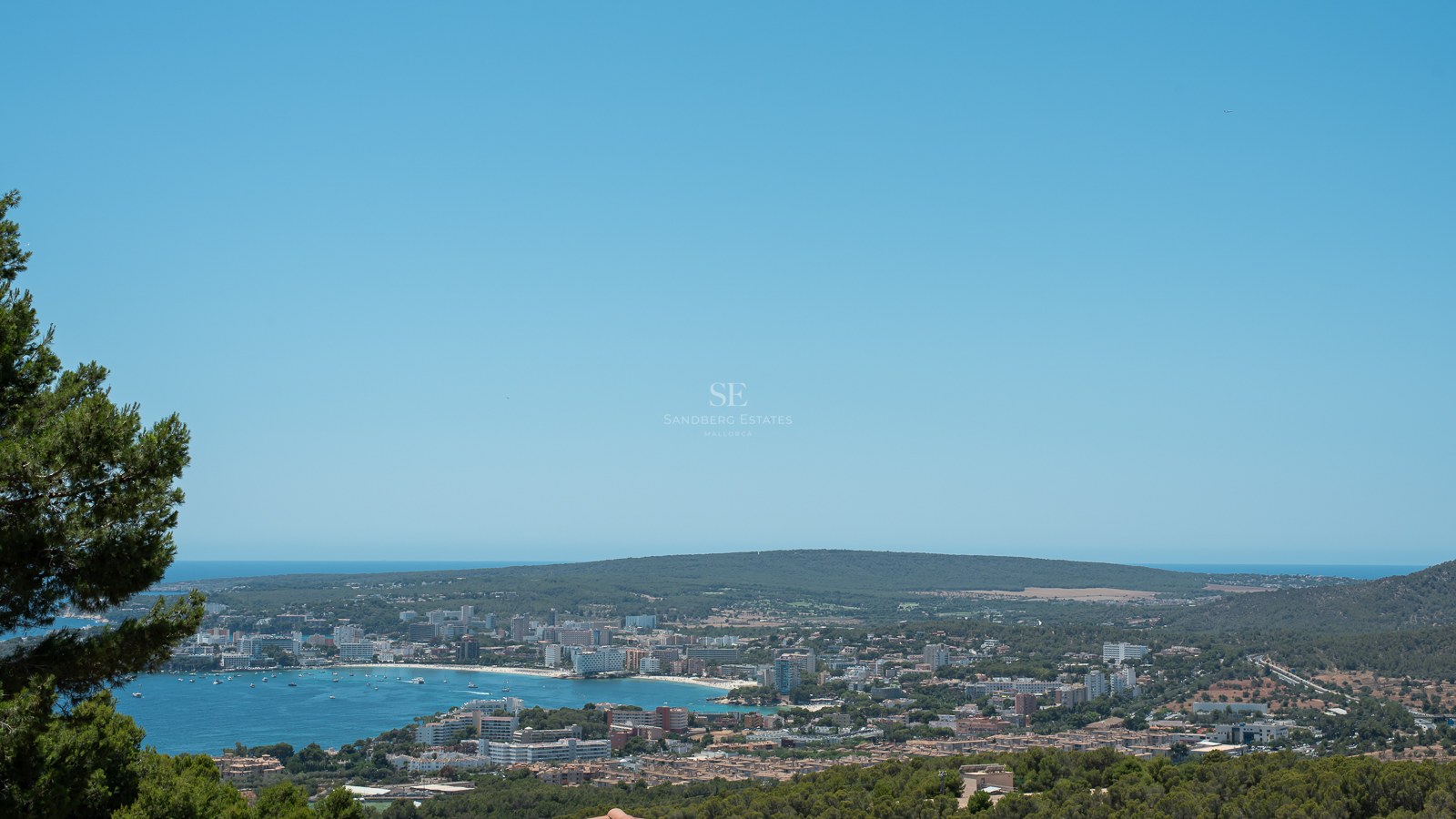 Vue panoramique surélevée d'une baie méditerranéenne bleue, d'une ville côtière et de collines verdoyantes.