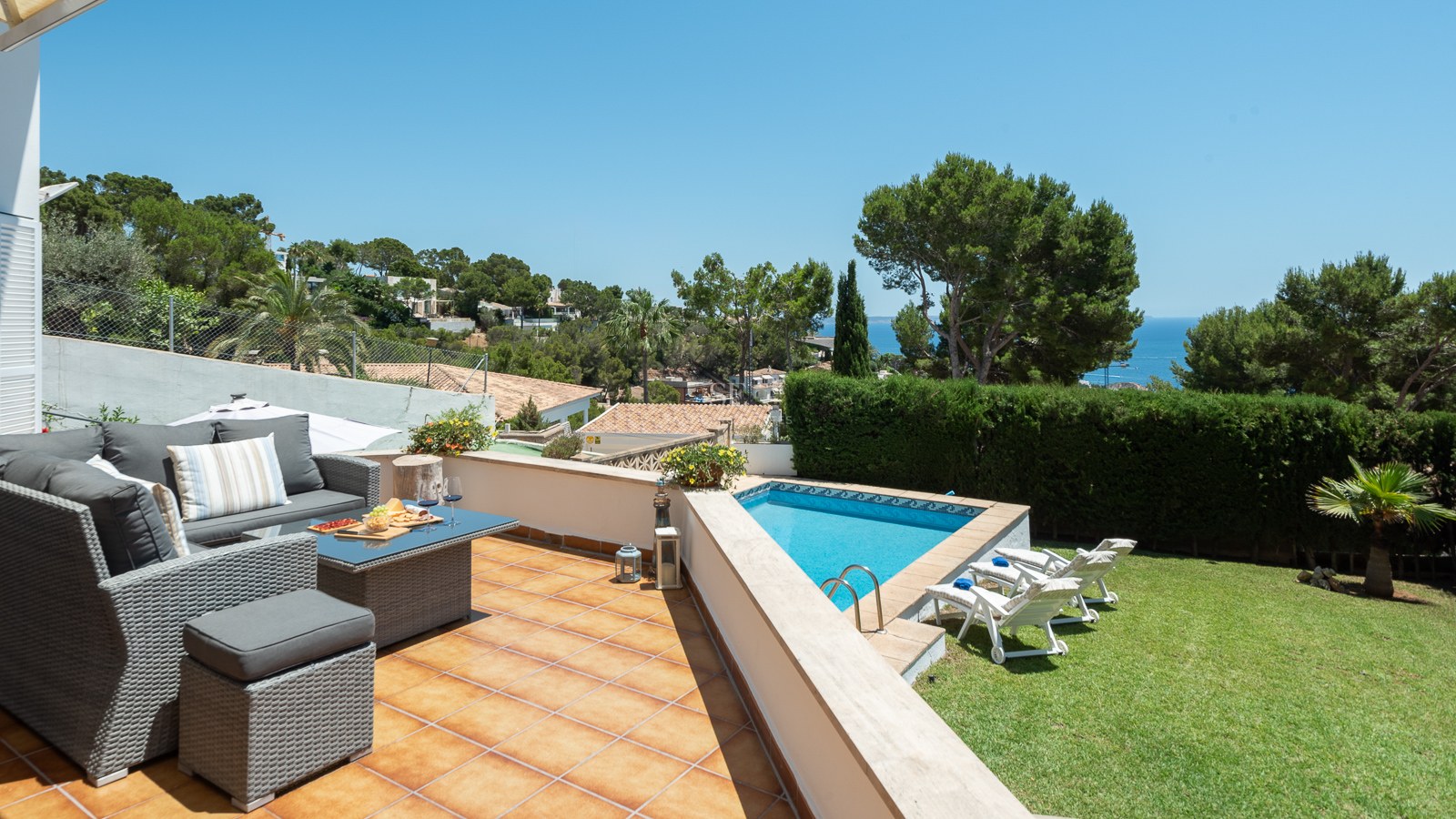 Terrasse ensoleillée avec canapés en résine tressée, piscine bleue, pelouse et vue panoramique sur l'océan.