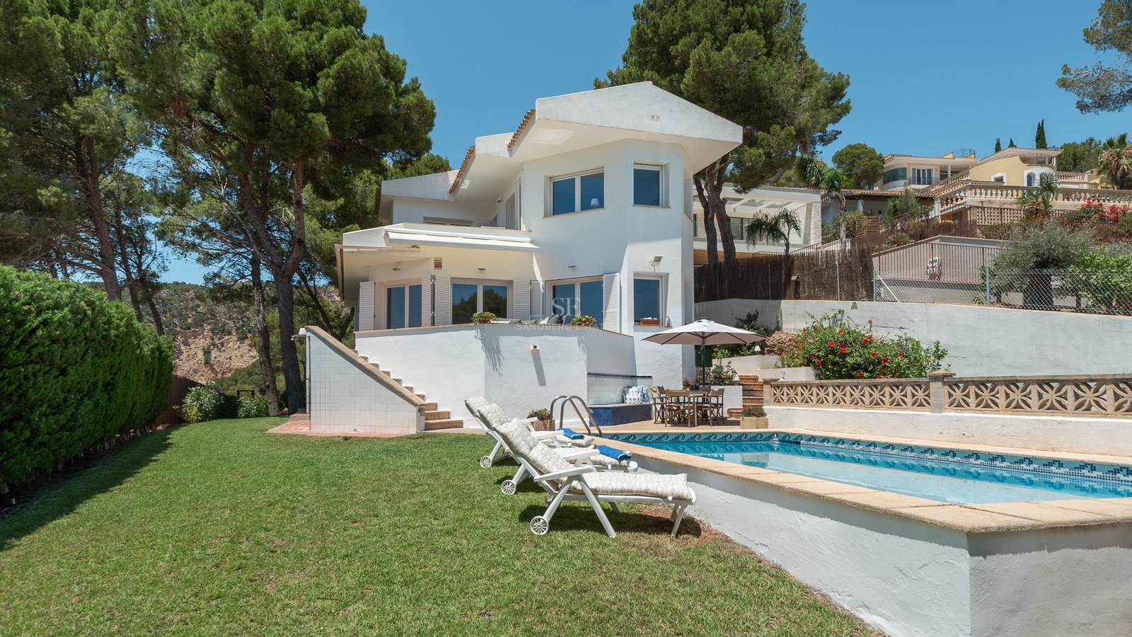 Villa blanche de deux étages avec piscine, transats et pelouse entourée de pins.