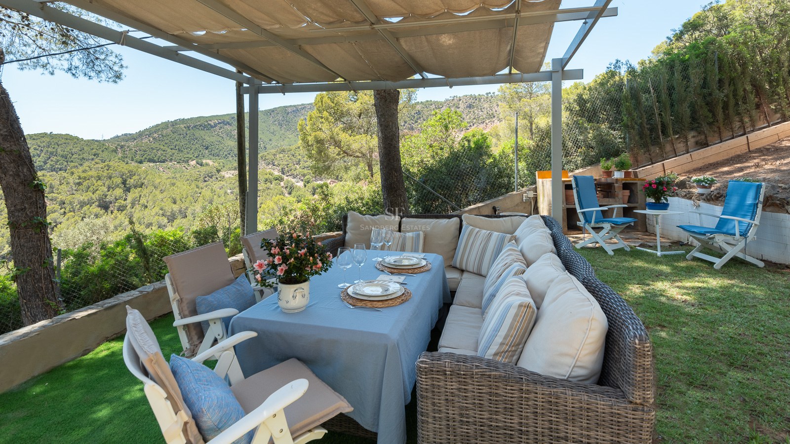 Espace dînatoire extérieur sous pergola avec canapés en osier et vue sur les collines verdoyantes.