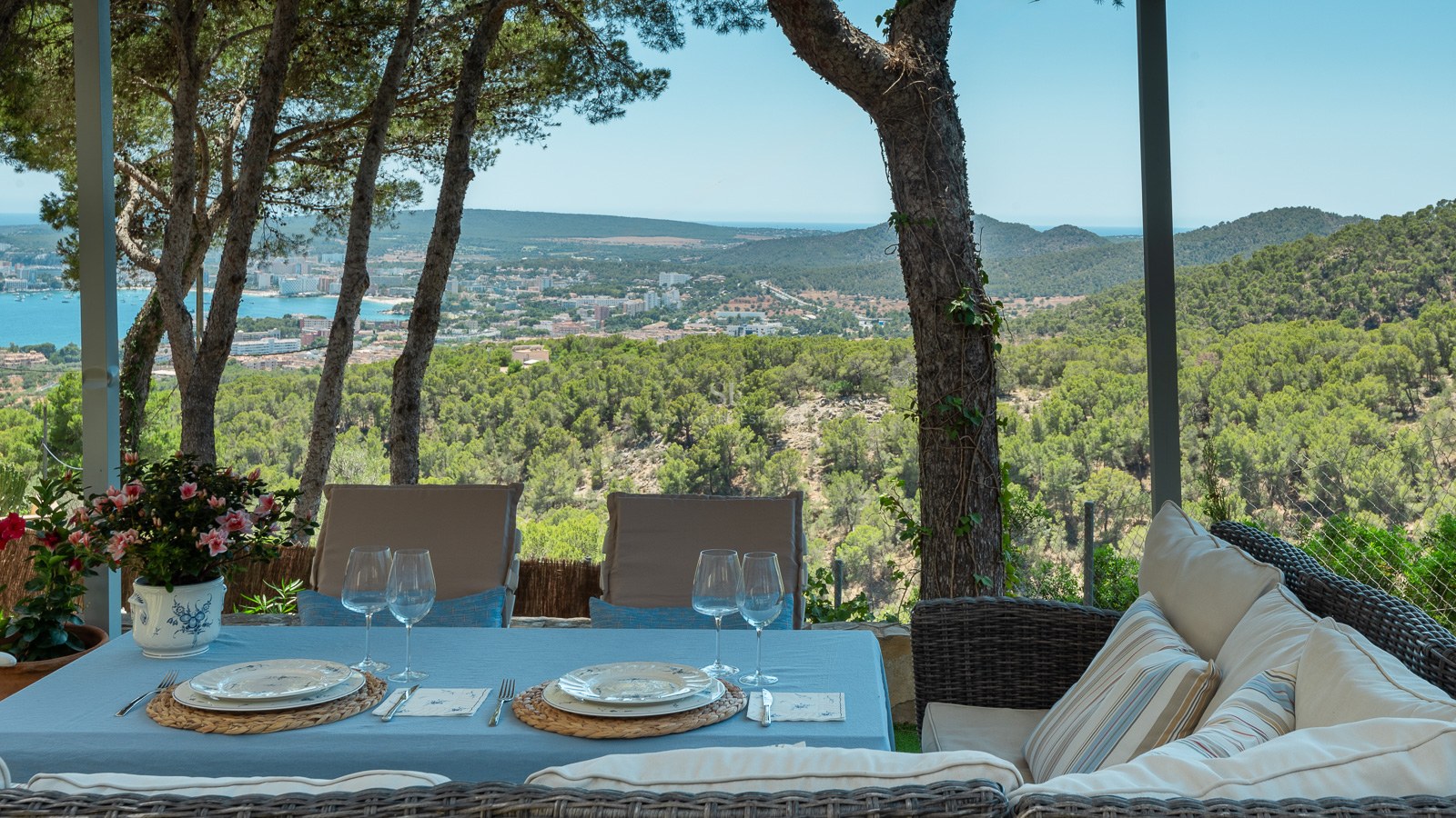 Table à manger extérieure et canapé en osier sur une terrasse ombragée surplombant une ville côtière et la mer.