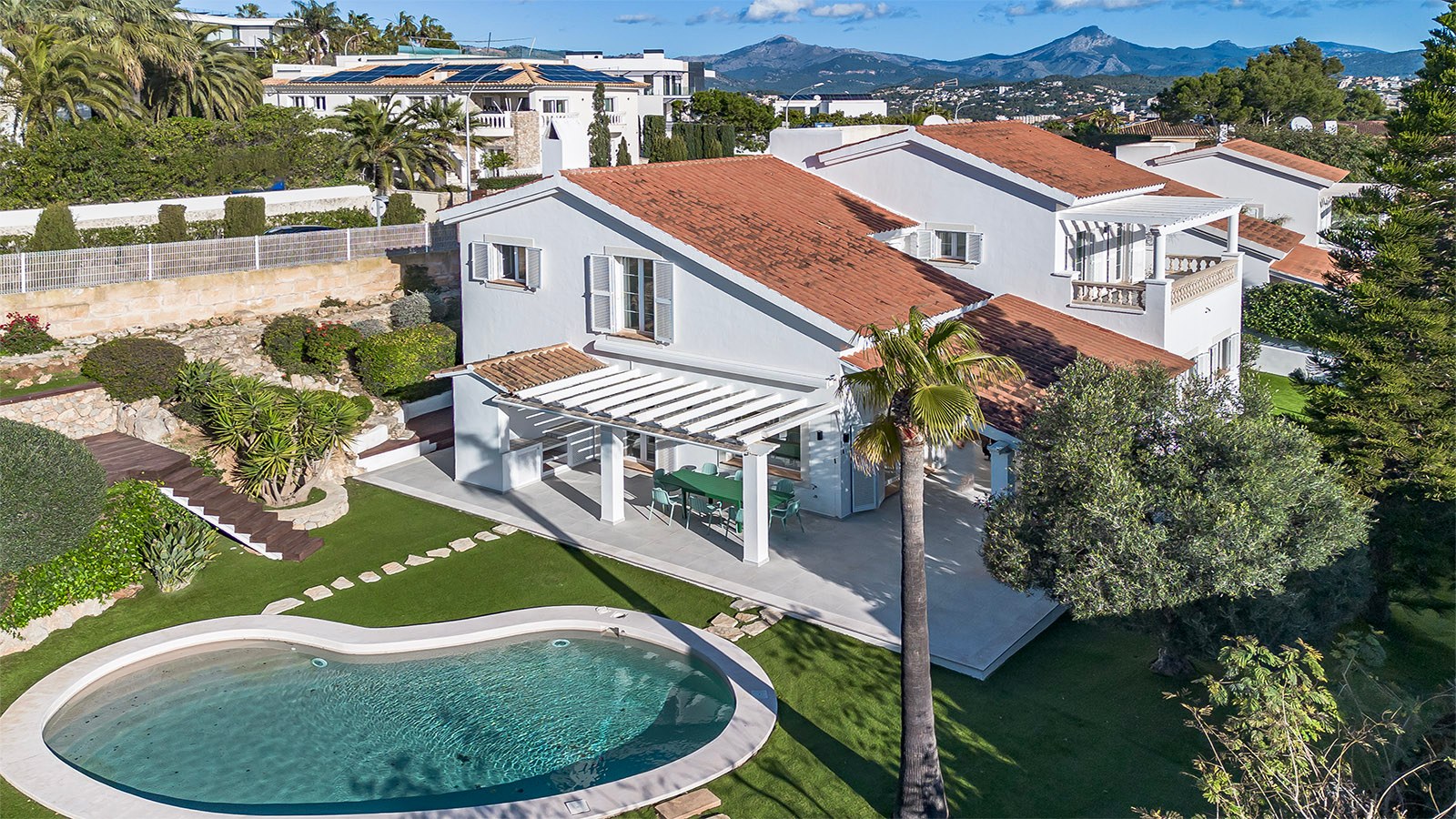 Vue aérienne d'une villa blanche, toit en terre cuite, piscine en forme de rein et jardin sur fond de montagnes.