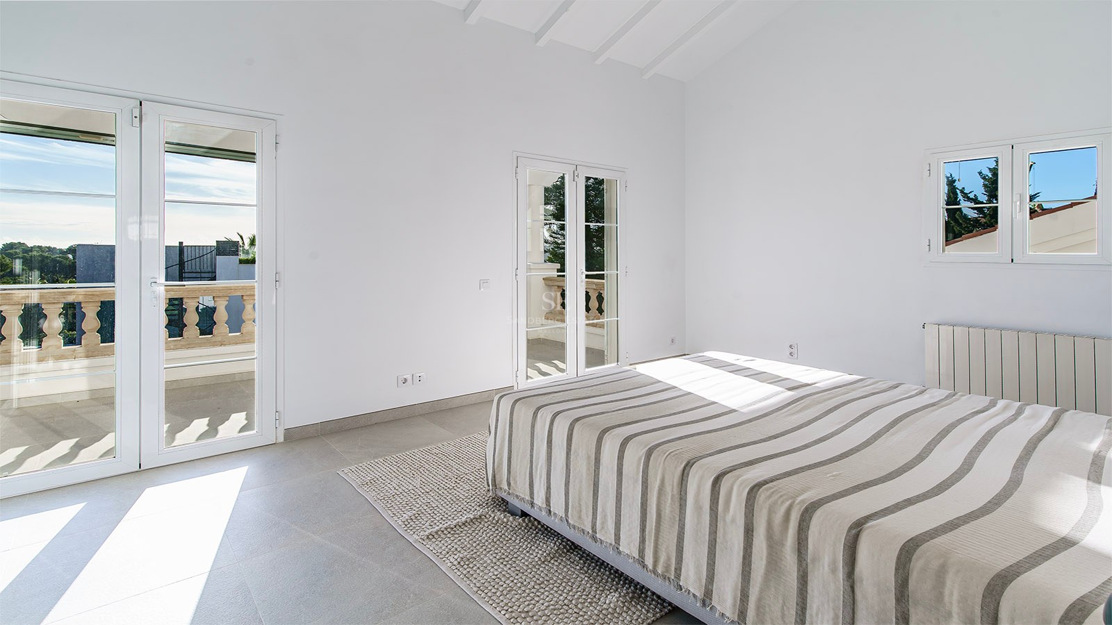Chambre minimaliste avec lit rayé, sol en grès gris, hauts plafonds à poutres blanches et balcon ensoleillé.