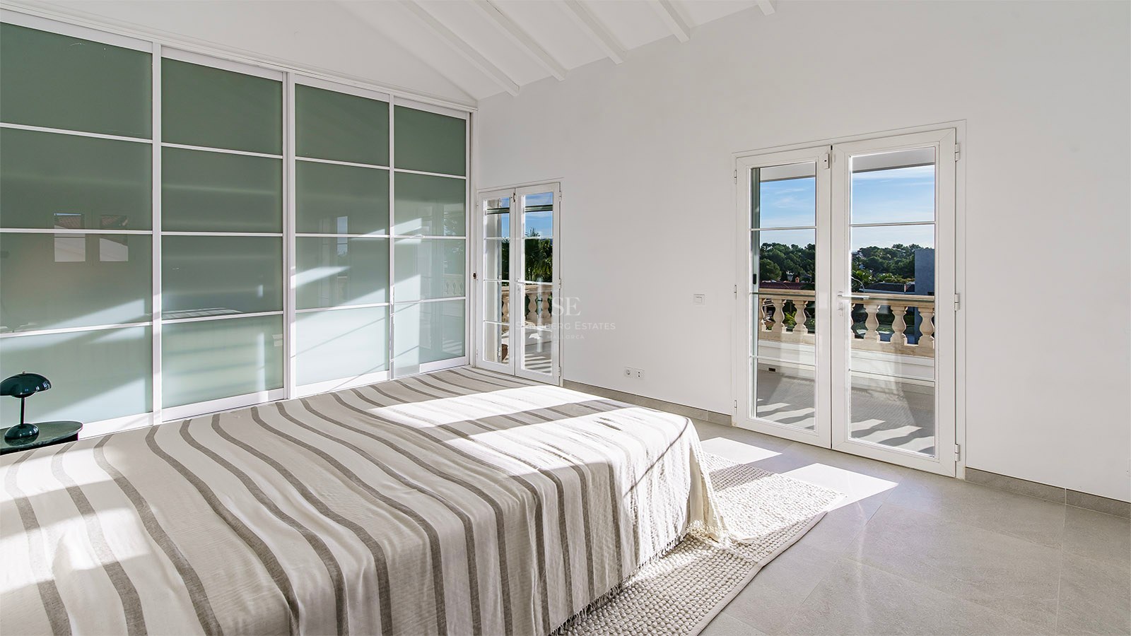 Chambre lumineuse avec lit rayé, plafonds voûtés blancs, placards en verre dépoli et accès à un balcon.