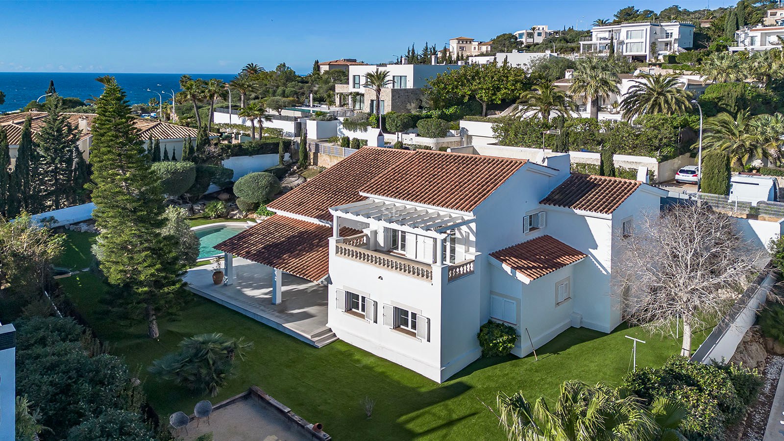 Vue aérienne d'une villa blanche méditerranéenne, toit en terre cuite, jardin, piscine et mer en arrière-plan.