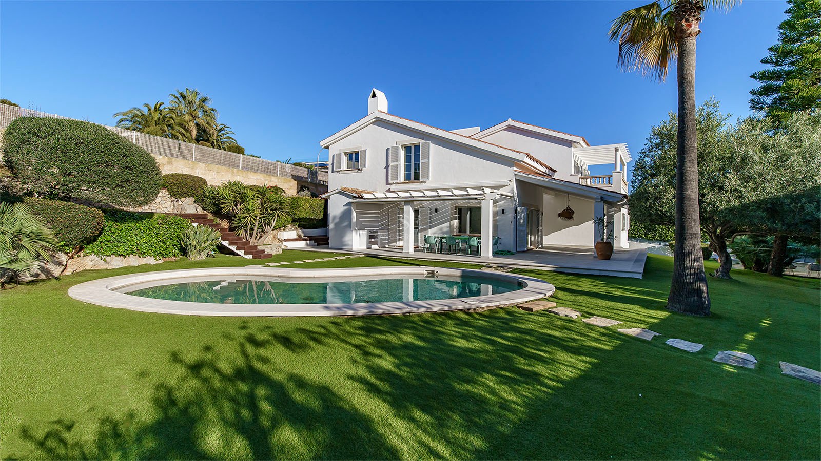 Villa blanche à deux étages avec piscine de forme organique, pelouse verte et palmiers sous un ciel bleu clair.