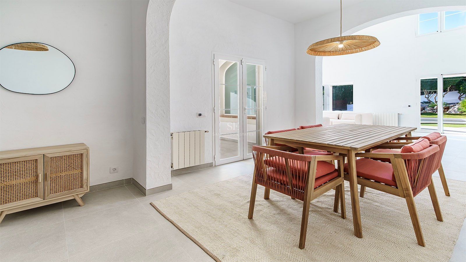 Salle à manger lumineuse avec table en bois, chaises terre cuite, luminaire en osier et arches blanches.