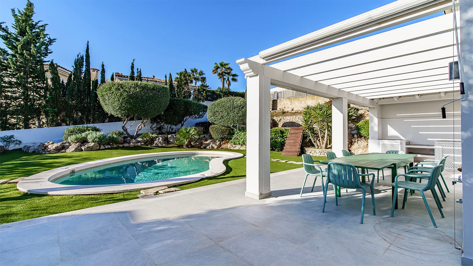 Pergola blanche abritant un coin repas extérieur avec chaises vertes à côté d'une piscine et d'un jardin paysager.