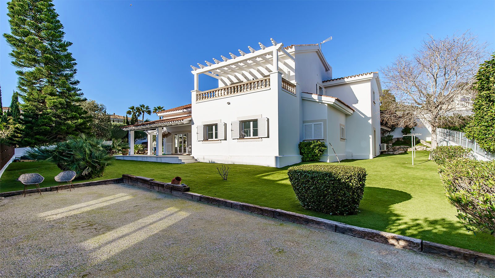 Villa blanche à deux étages avec pergola, pelouse soignée et pins parasols sous un ciel bleu intense.