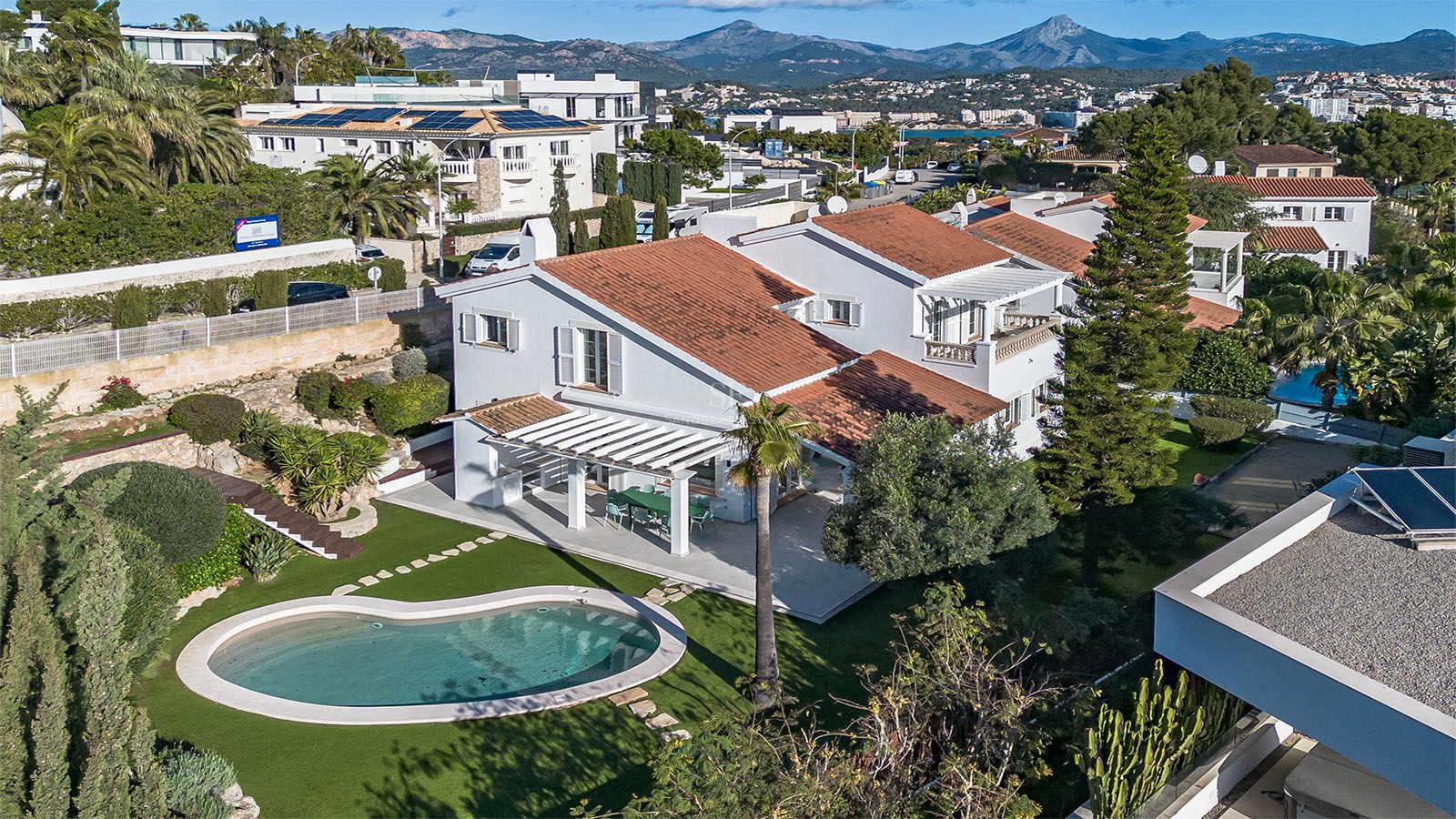 Vue aérienne d'une villa blanche au toit de tuiles avec piscine en forme de haricot et jardin devant les montagnes.