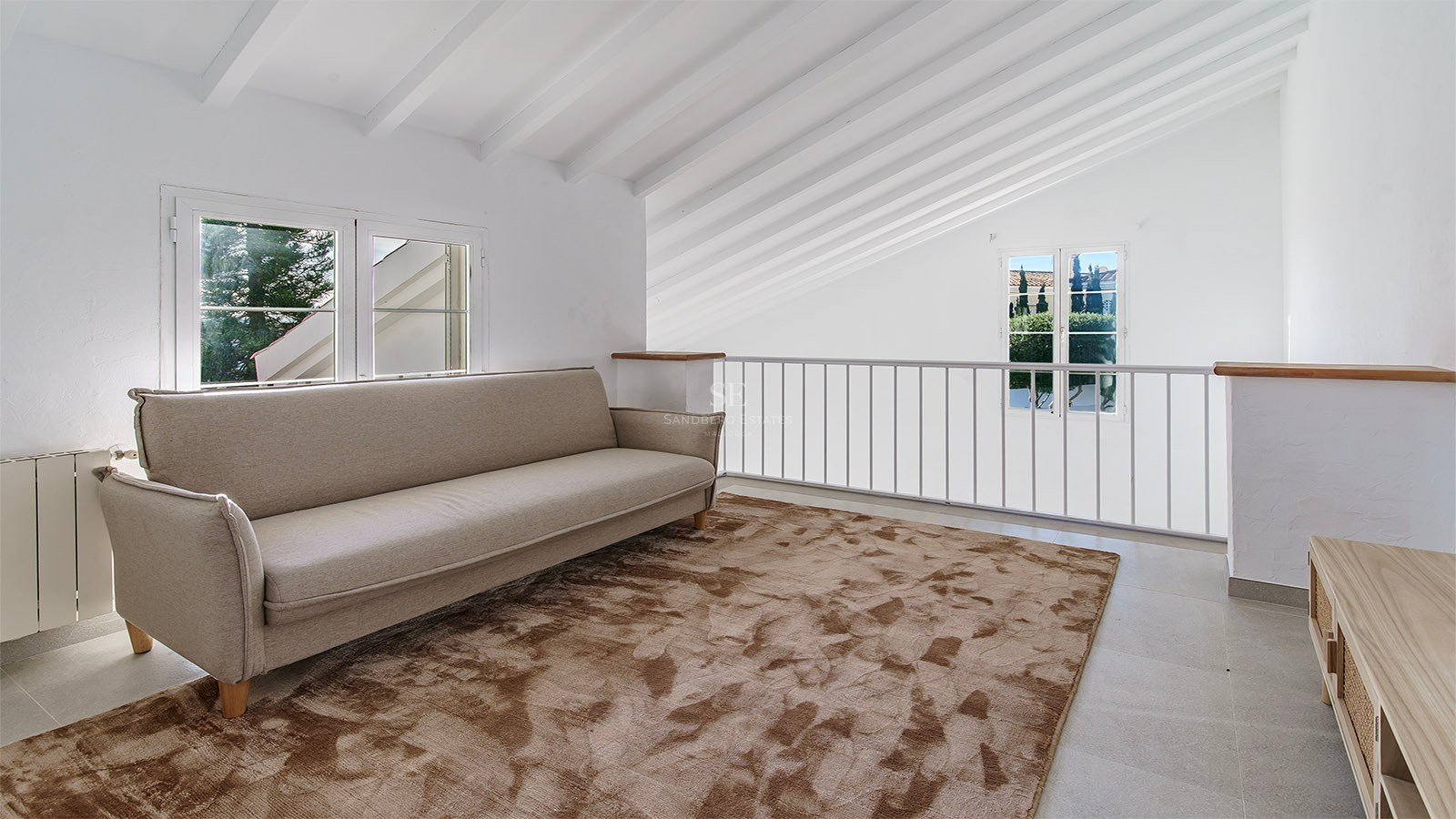 Salon en mezzanine avec canapé beige, tapis brun texturé et plafond mansardé avec poutres en bois blanc.