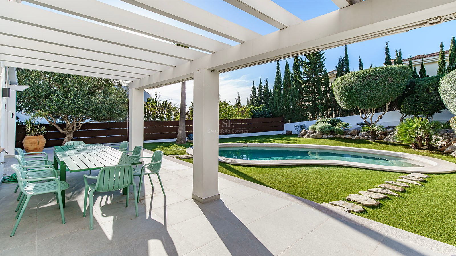 Terrasse ombragée avec coin repas vert donnant sur une piscine de forme libre et un jardin paysager avec cyprès.