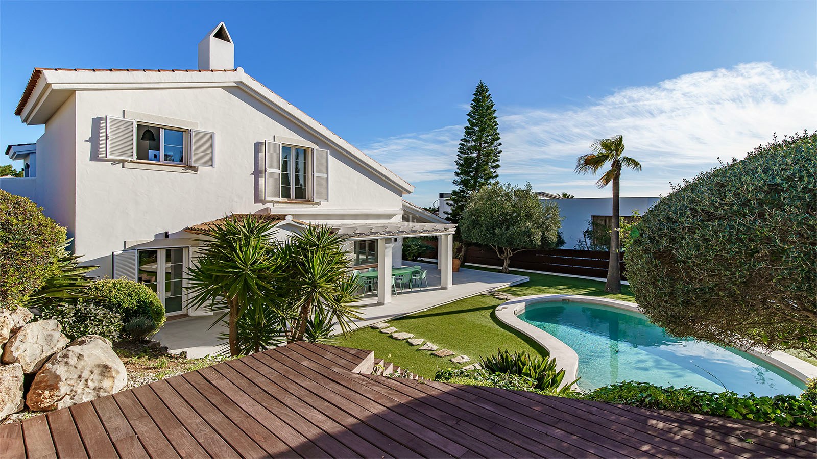 Villa blanche à deux étages avec une piscine en forme de haricot, une terrasse en bois et un jardin arboré.