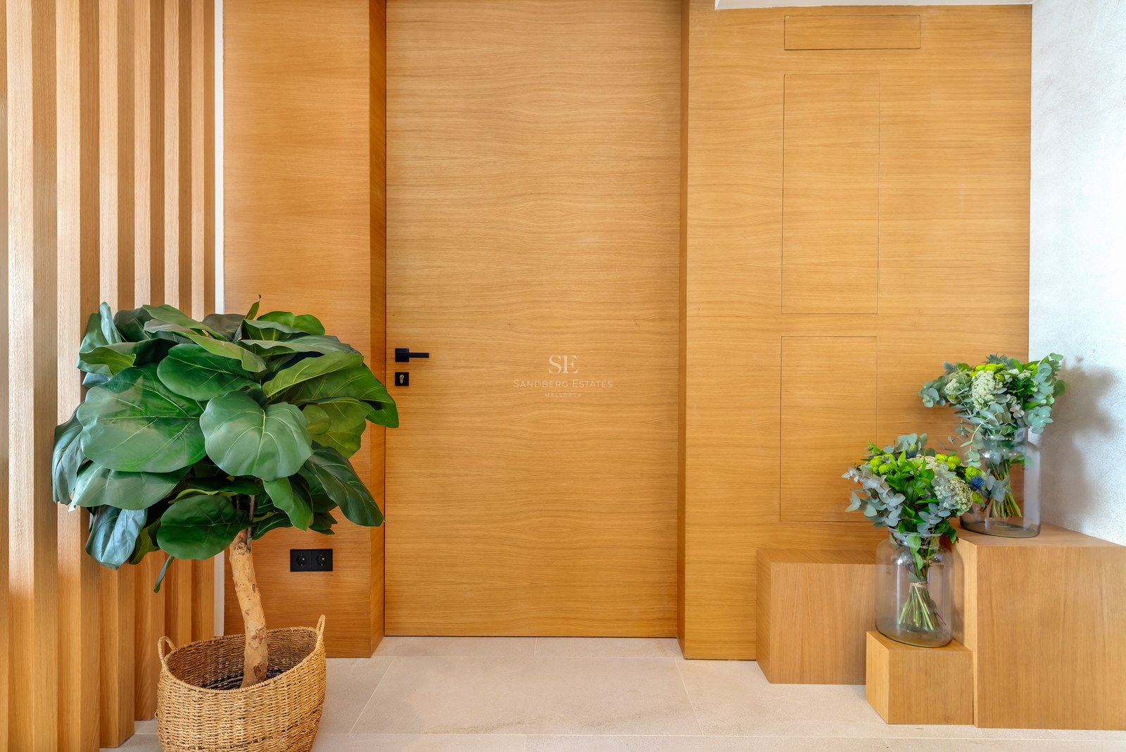 Puerta de roble claro de suelo a techo con paneles de pared a juego, manivela negra y planta en cesta de mimbre.