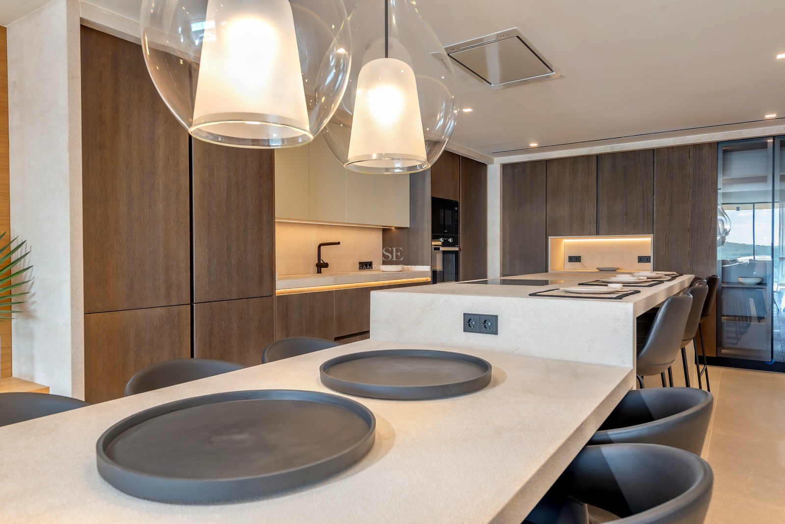 Cocina moderna con armarios de madera oscura, isla de piedra clara con taburetes y lámparas colgantes de cristal.