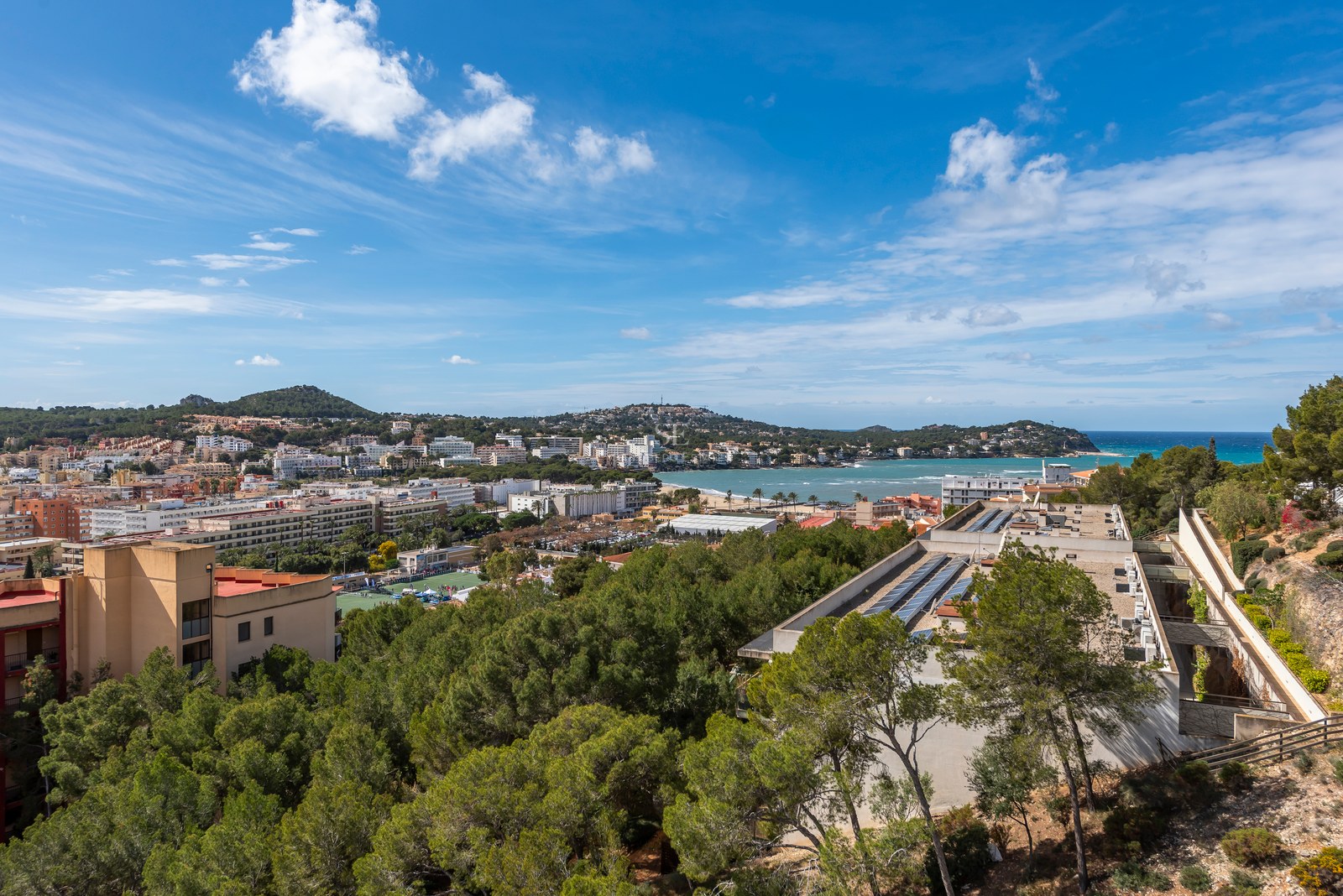 Vista elevada de un pueblo costero mediterráneo con mar azul, bosques de pinos y montañas bajo un cielo despejado.
