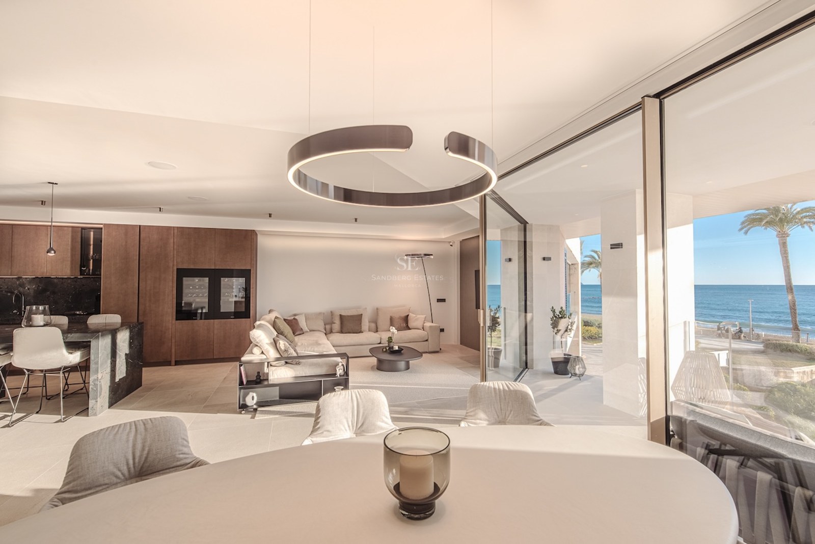 Bright open-plan living room with floor-to-ceiling glass doors opening to a terrace with sea views and palm trees.