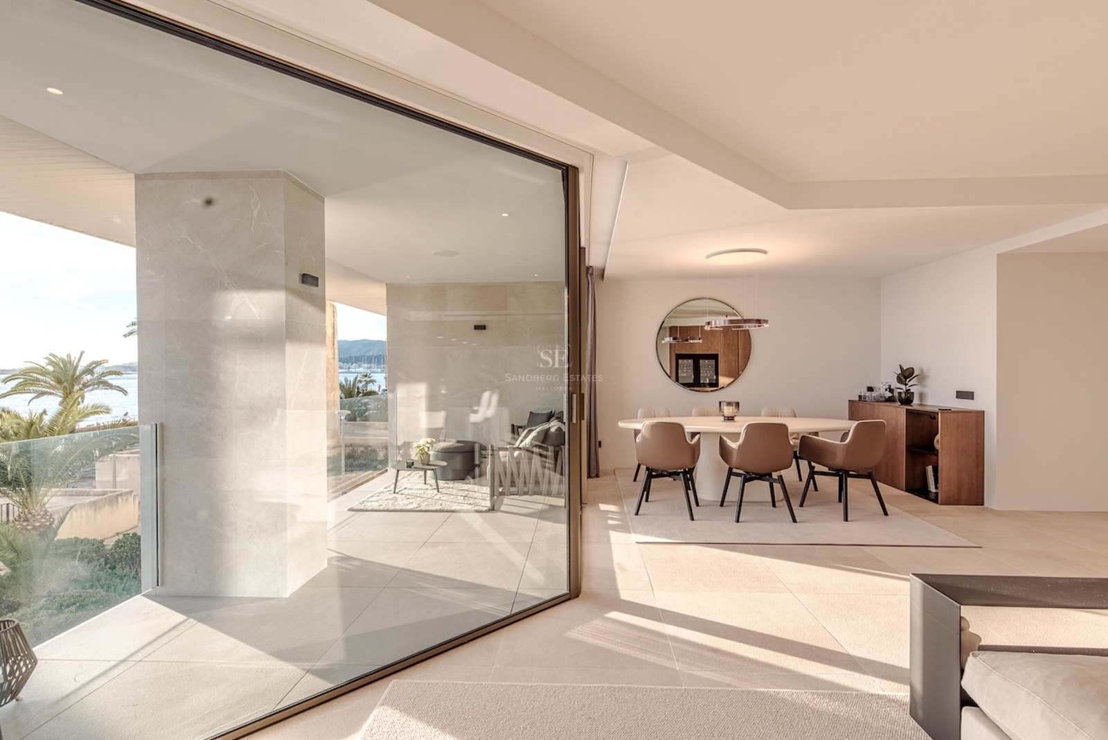 Interior dining room with round table and brown chairs, opening through glass doors to a sunny terrace with sea views.
