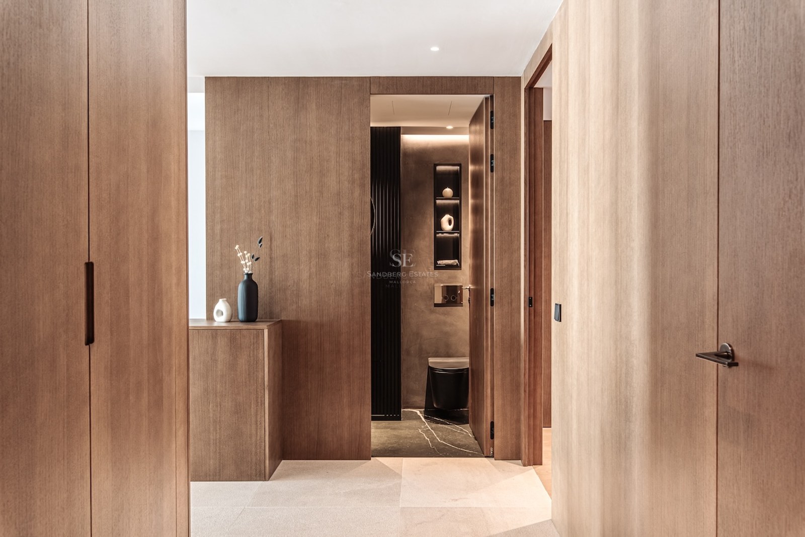 Hallway featuring floor-to-ceiling oak cabinetry leading into a minimalist modern bathroom with dark finishes.
