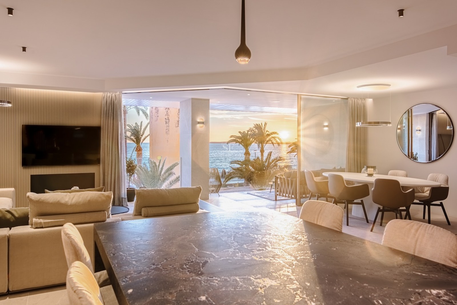 Luxury open-plan living and dining room with floor-to-ceiling glass doors opening onto a terrace with sea views at sunset.