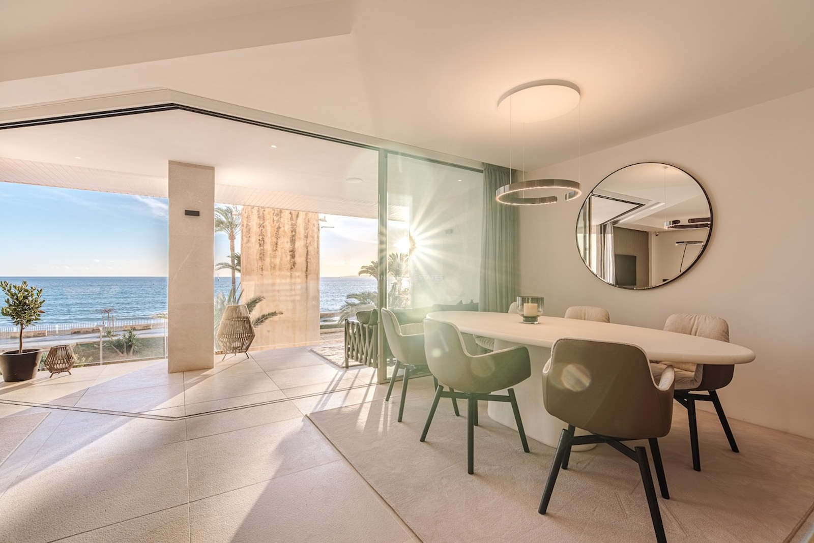 Modern indoor dining area with an oval table and floor-to-ceiling windows opening to a sunlit terrace by the sea.