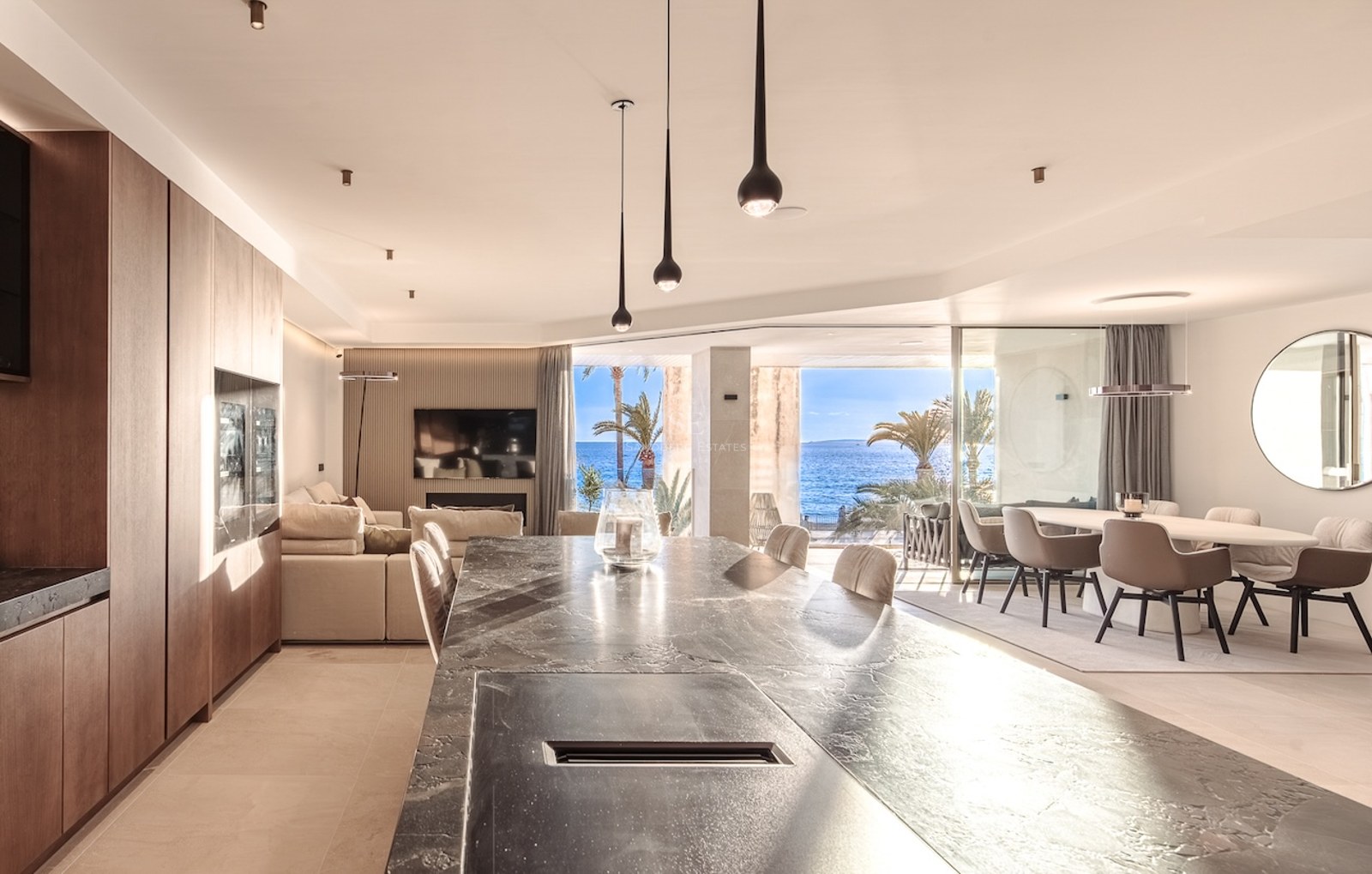 Modern open-plan kitchen and living room featuring a dark stone island, wood cabinetry, and floor-to-ceiling windows overlooking the sea.