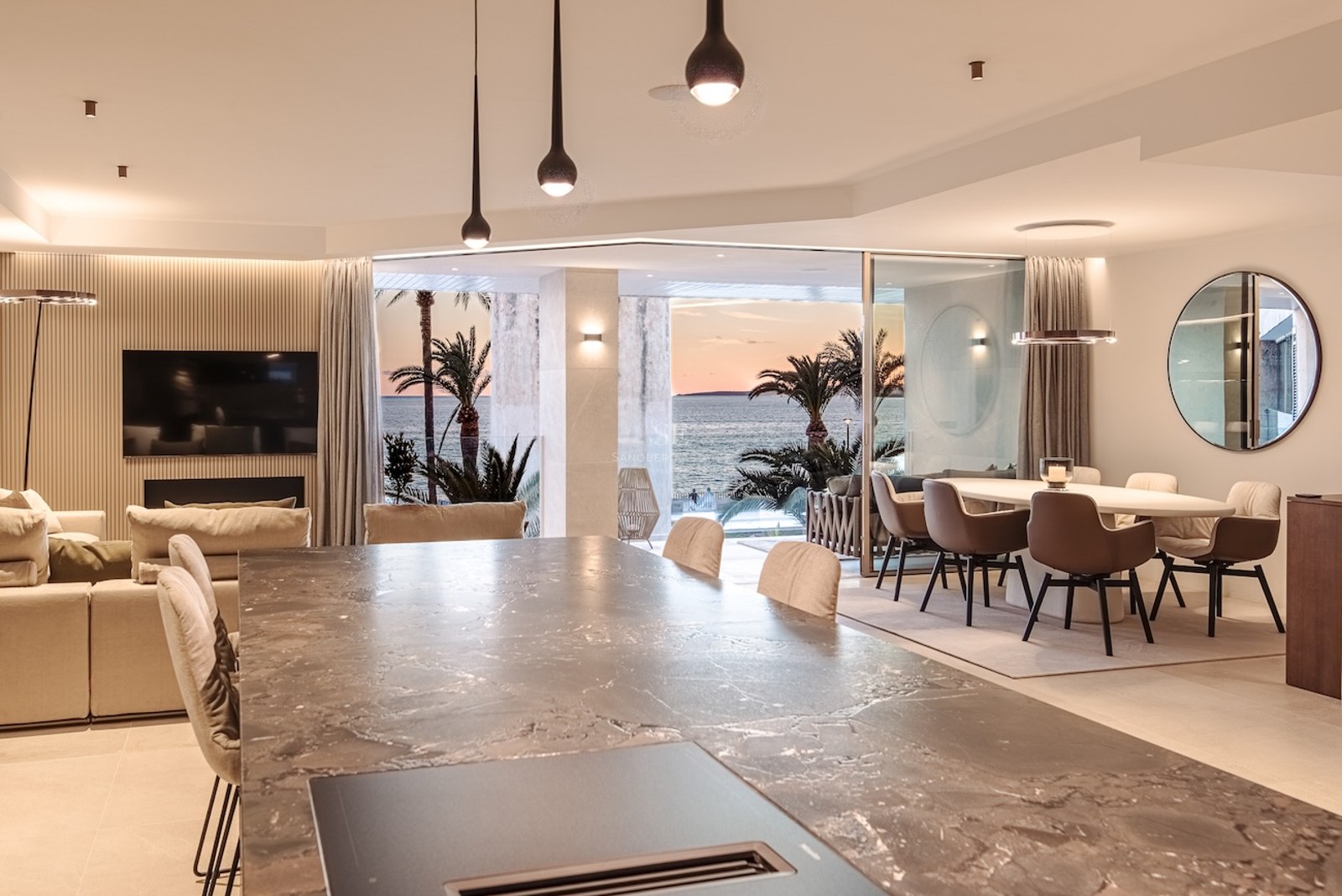 Modern open-plan living and dining space with a marble island, overlooking a terrace with palms and the sea at sunset.
