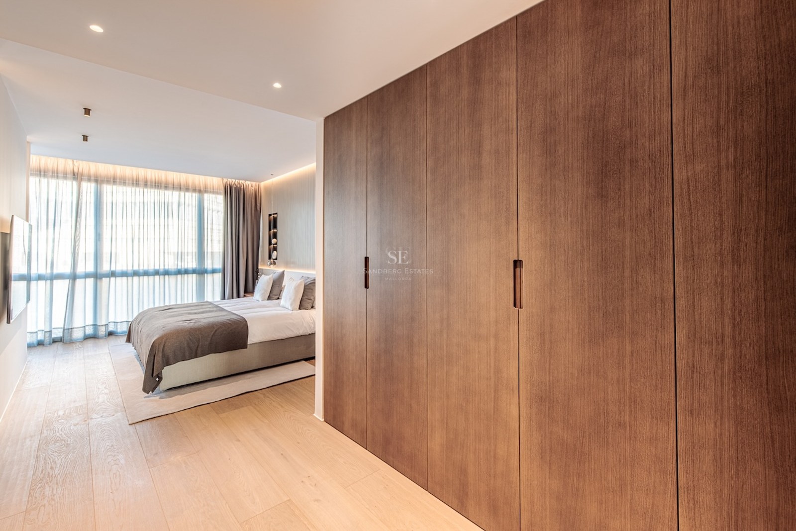 Modern bedroom featuring floor-to-ceiling wood wardrobes, light oak flooring, and large windows with soft curtains.