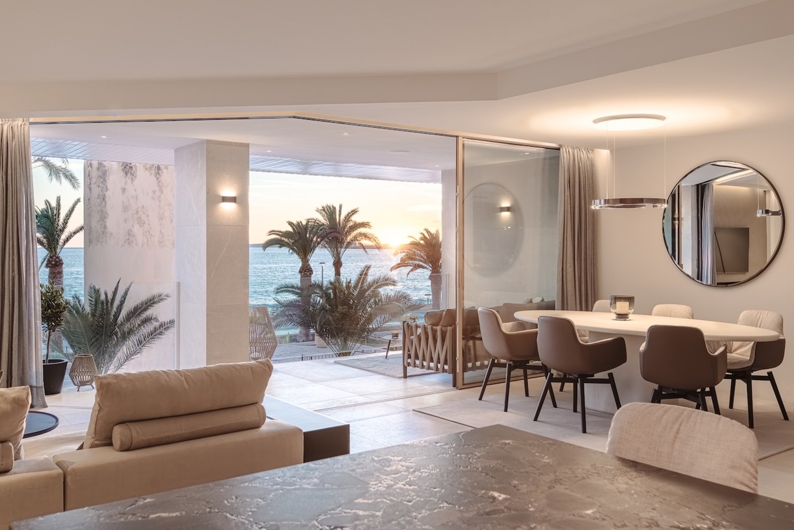 Contemporary dining room with marble table opening onto a terrace with palm trees and sunset sea views.