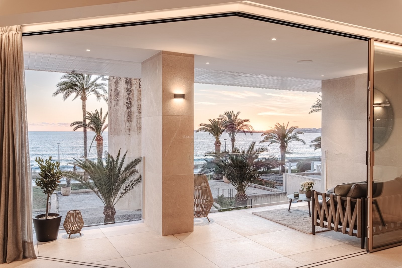 Modern terrace with stone flooring and glass railings overlooking the sea and palm trees at sunset.
