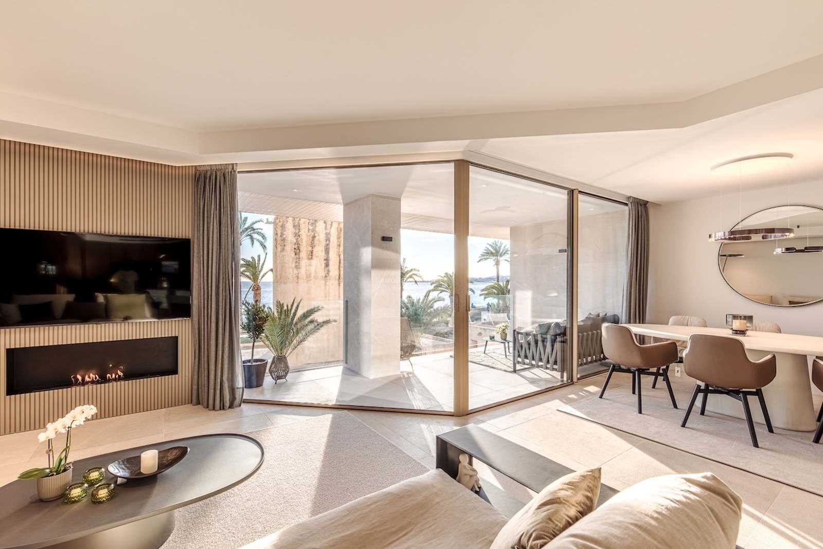 Modern living and dining room with glass doors opening to a terrace with sea views and a slatted wood fireplace wall.