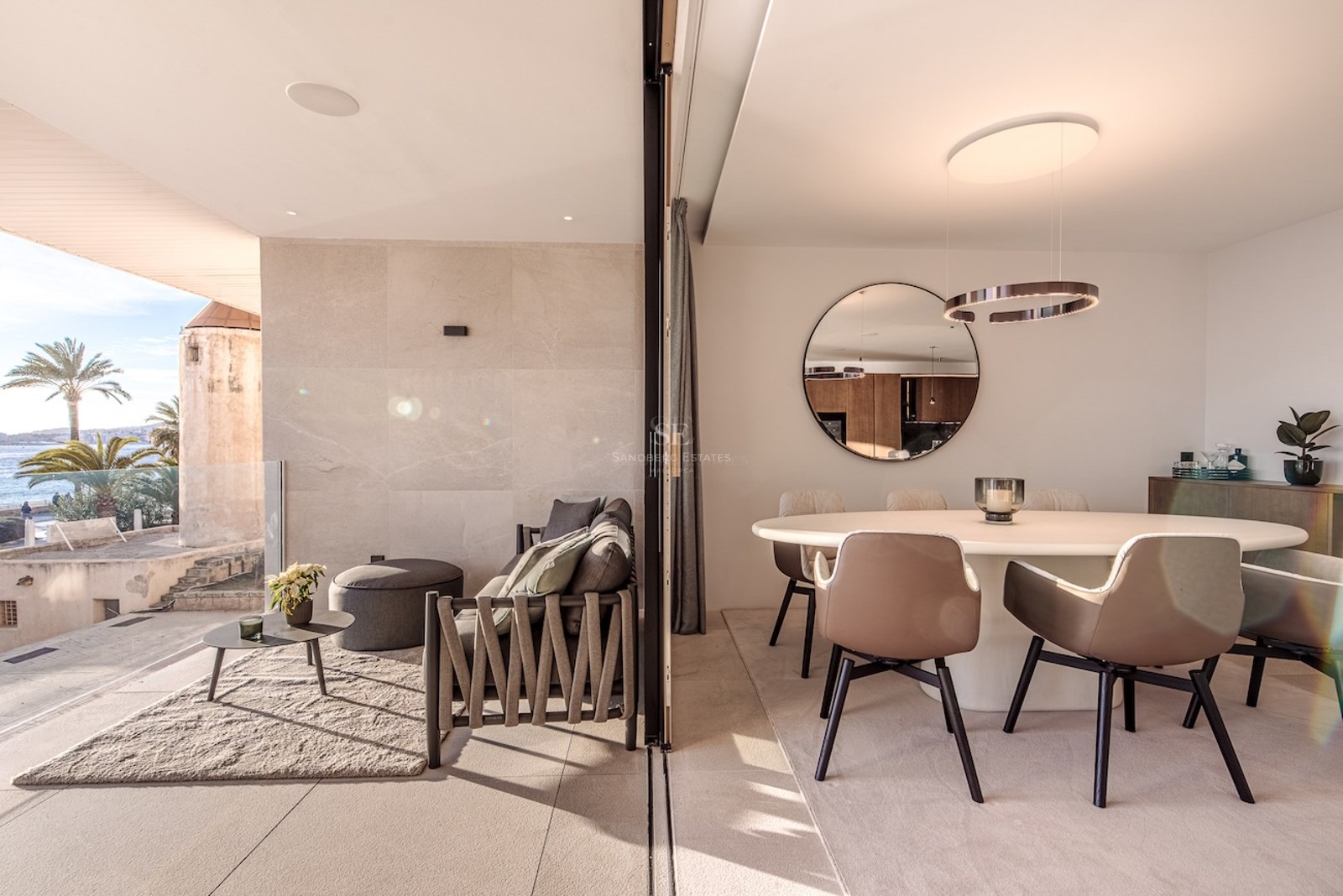 A modern dining room with a white round table and beige chairs, transitioning into an outdoor terrace with sea views.