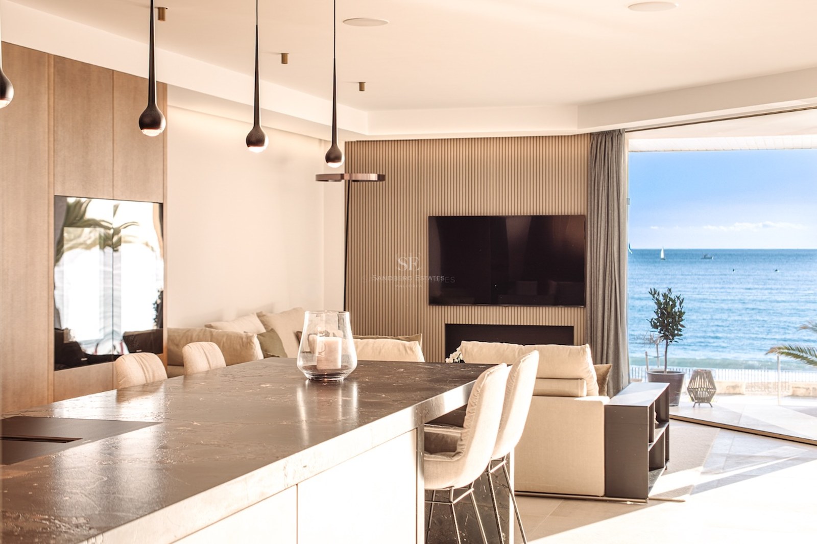Luxury open-plan living room and kitchen featuring a dark stone island and floor-to-ceiling windows overlooking the sea.