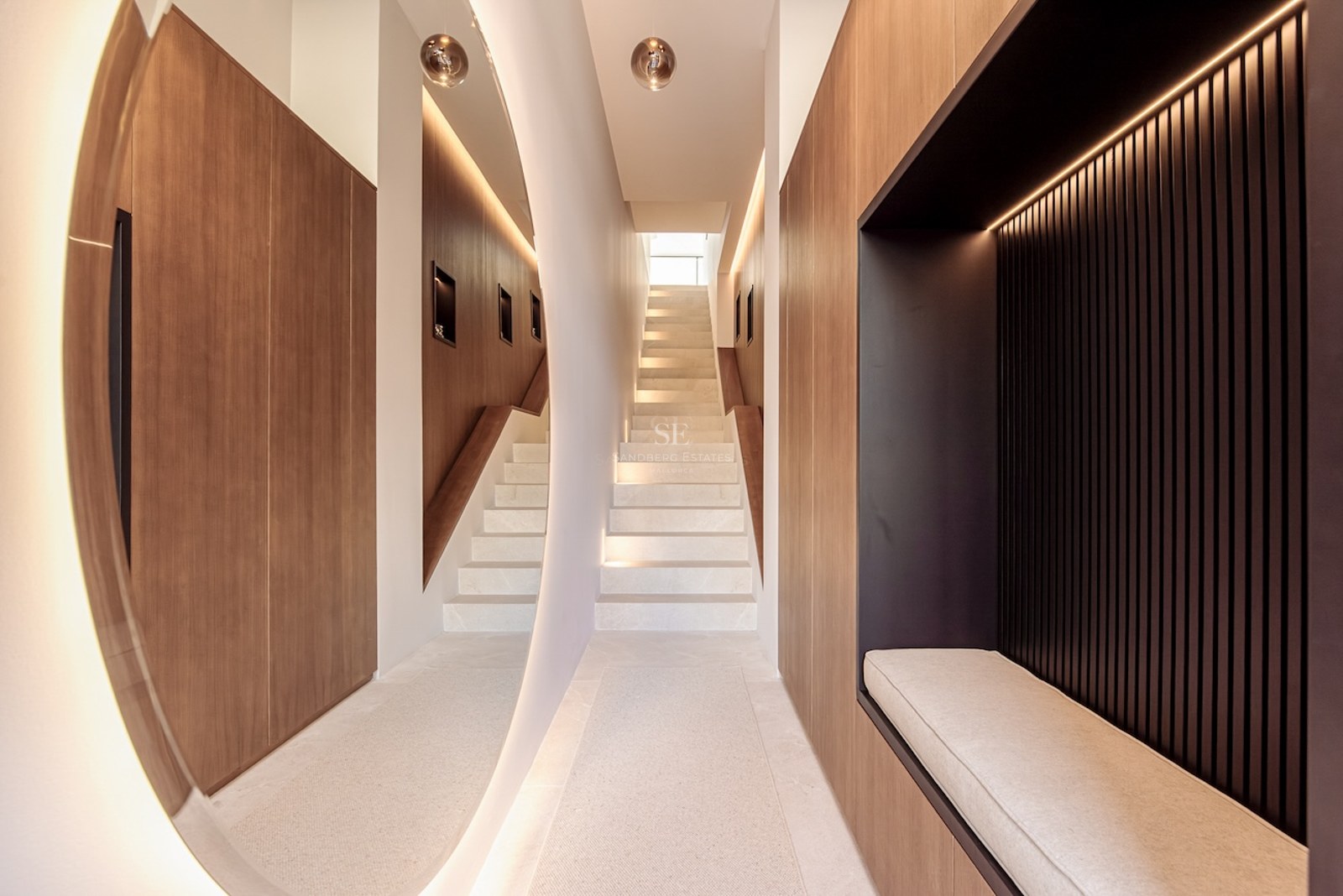 Modern hallway with floating stone staircase, wood paneling, large backlit circular mirror, and built-in seating niche.