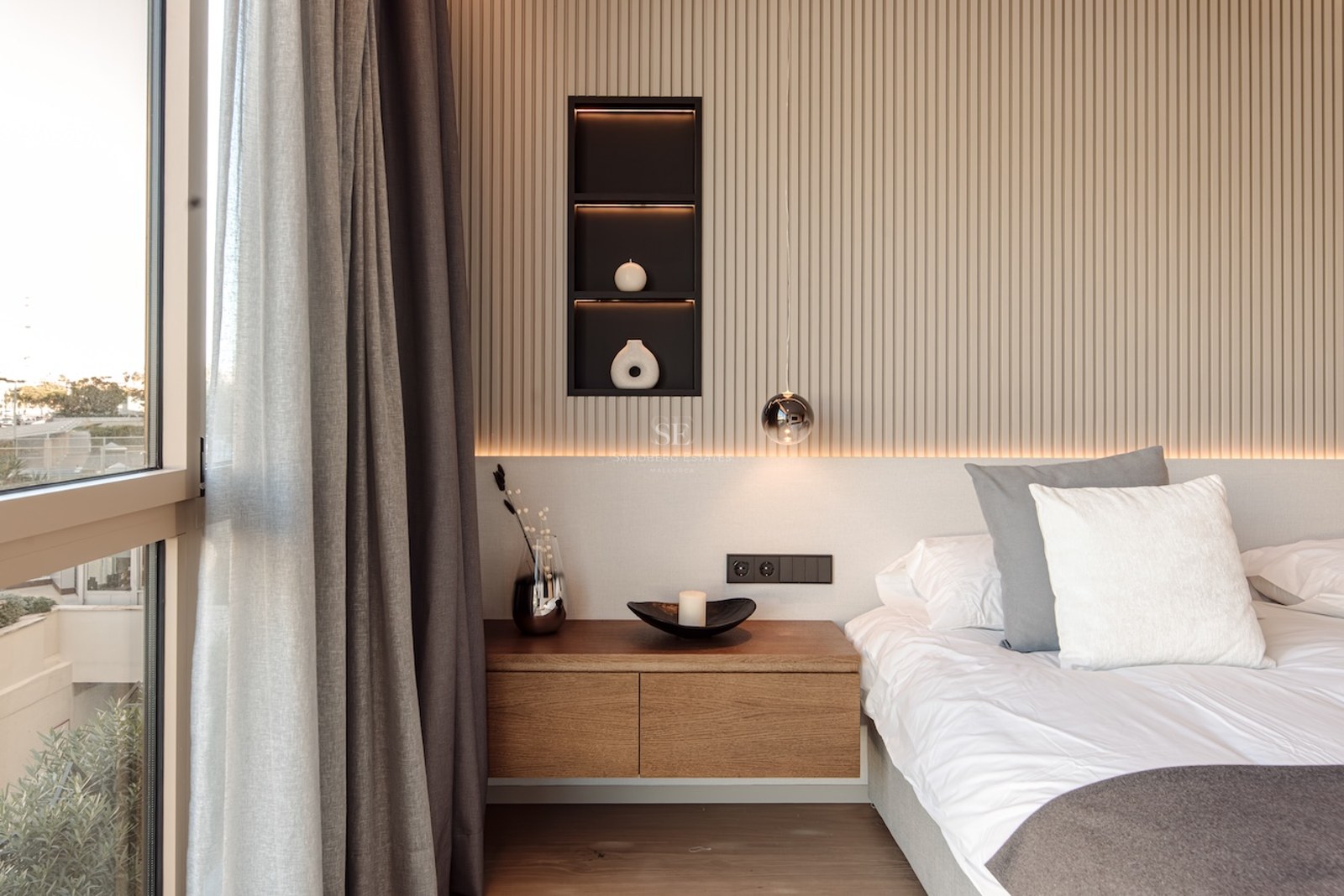 Modern bedroom featuring a slatted wood wall, floating oak nightstand, and large window with grey curtains.