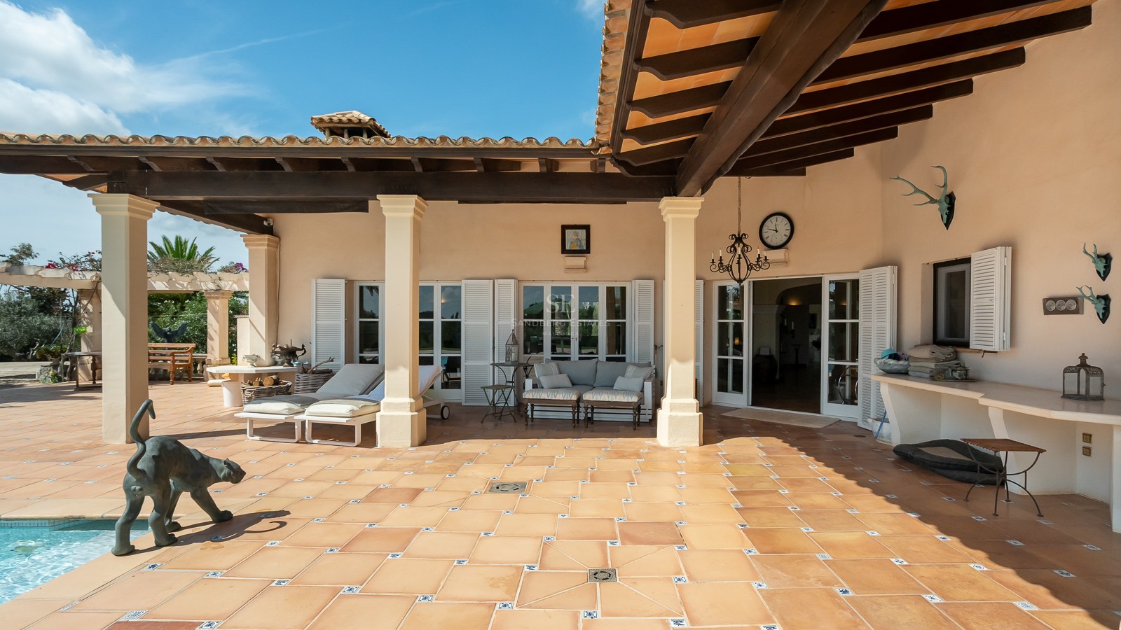 Terrakotta-Terrasse mit dunklen Holzbalken, weißen Säulen, Sonnenliegen und weißen Fensterläden im Sonnenlicht.