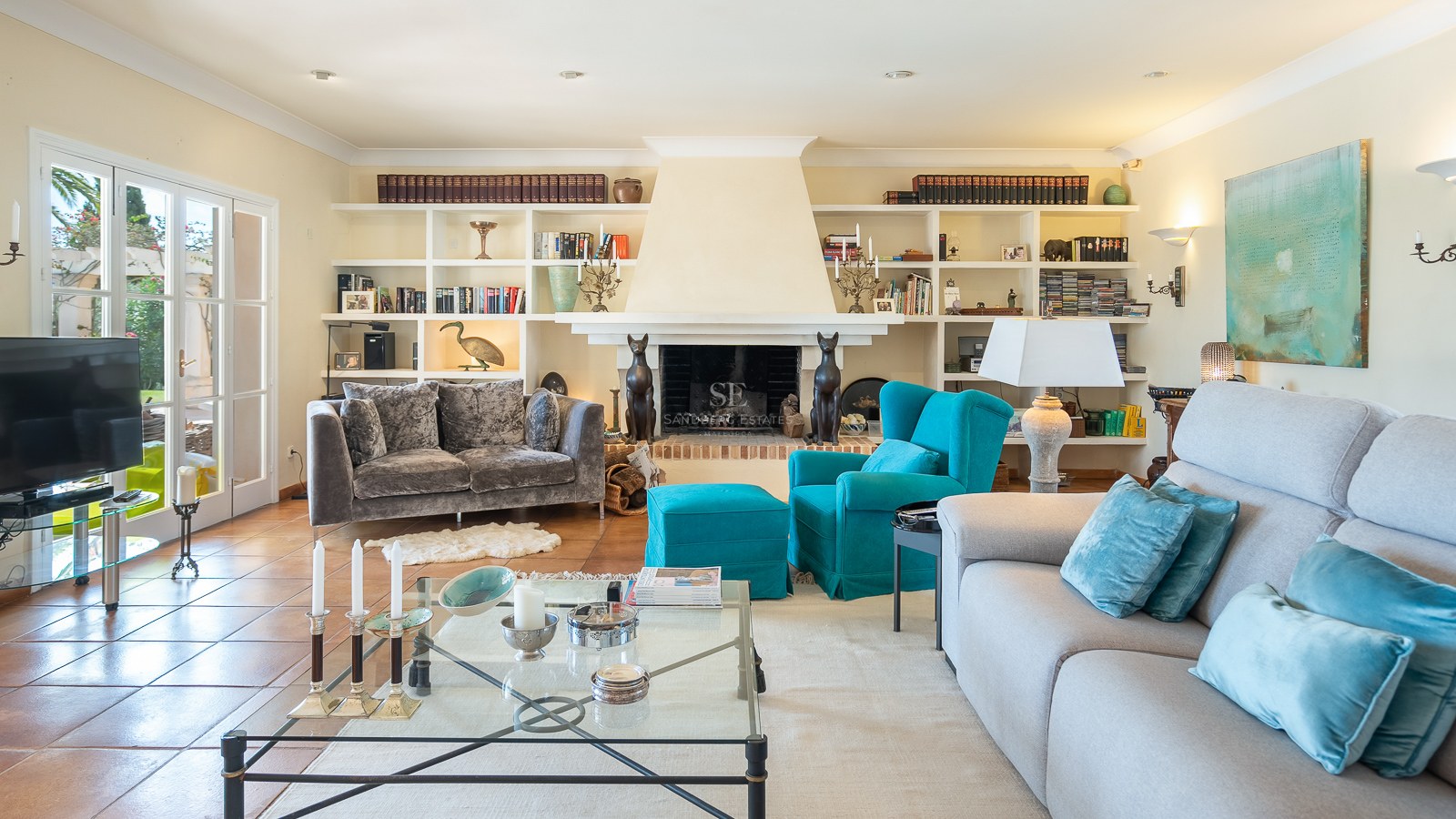 Helles Wohnzimmer mit Terrakottaboden, grauen Sofas, türkisfarbenem Sessel, Kamin und Wandregalen.