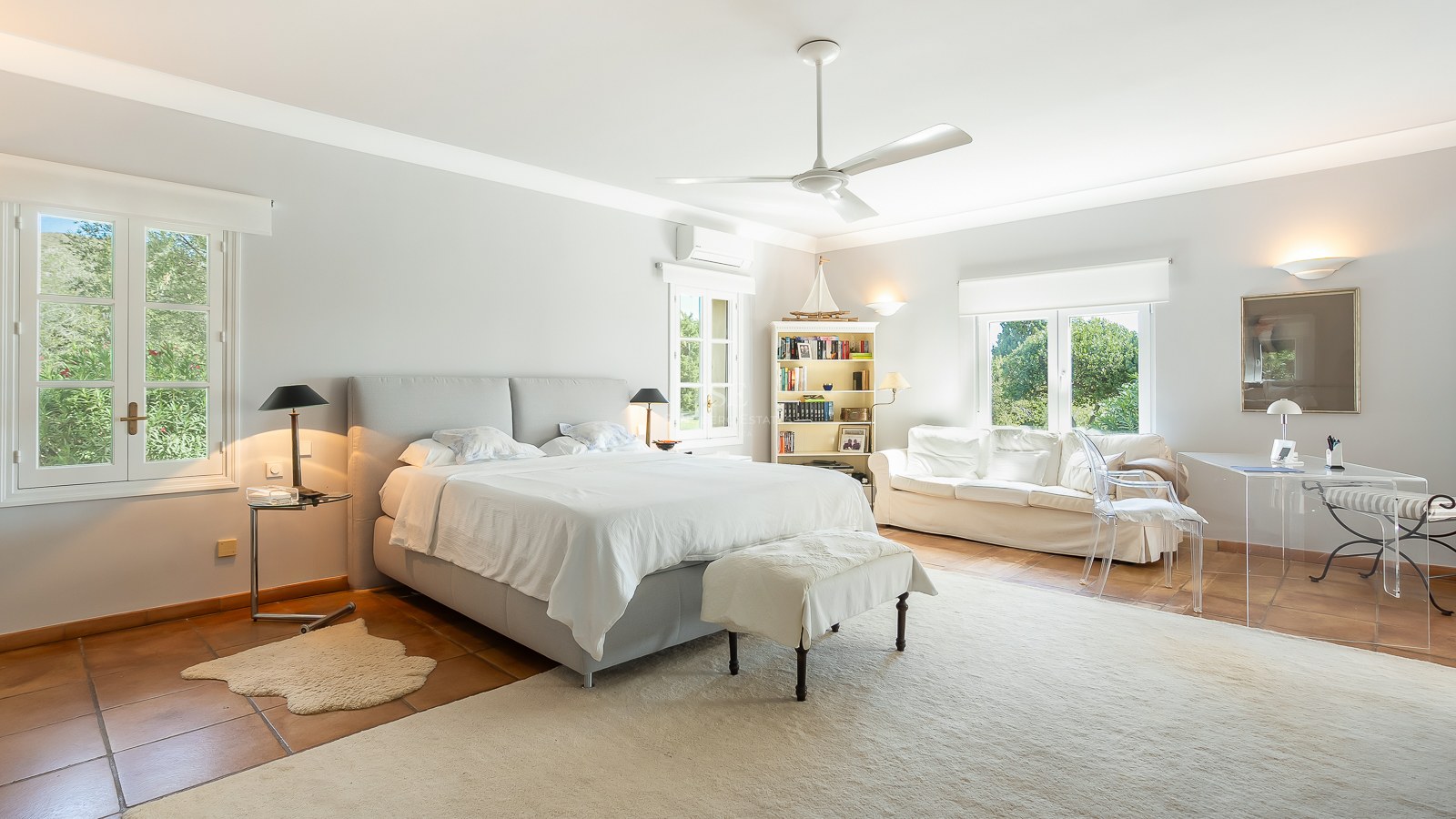 Großzügiges Hauptschlafzimmer mit Terrakottaboden, weißem Sofa, Glastisch und Fenstern mit Blick ins Grüne.