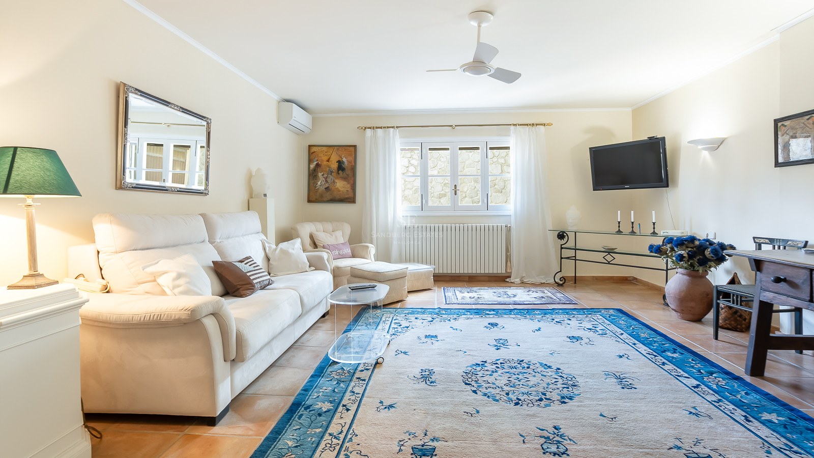 Helles Wohnzimmer mit weißem Sofa, großem blauem Teppich, Terrakottaboden und Deckenventilator.