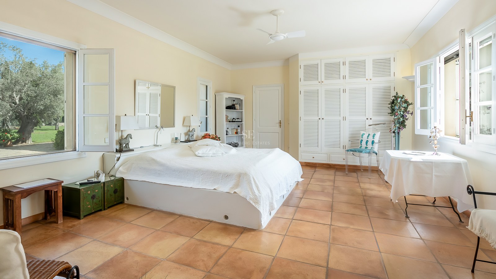 Geräumiges Schlafzimmer mit Terrakottaboden, weißen Lamellenschränken und Fenstern mit Blick auf einen grünen Garten.