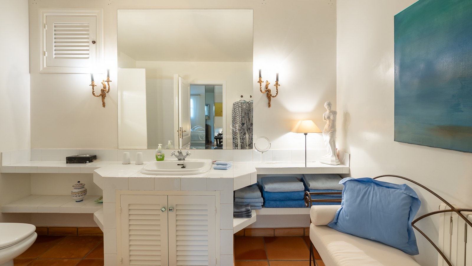 Salle de bain carrelée en blanc avec sol en terre cuite, grand miroir, appliques dorées et touches de bleu.