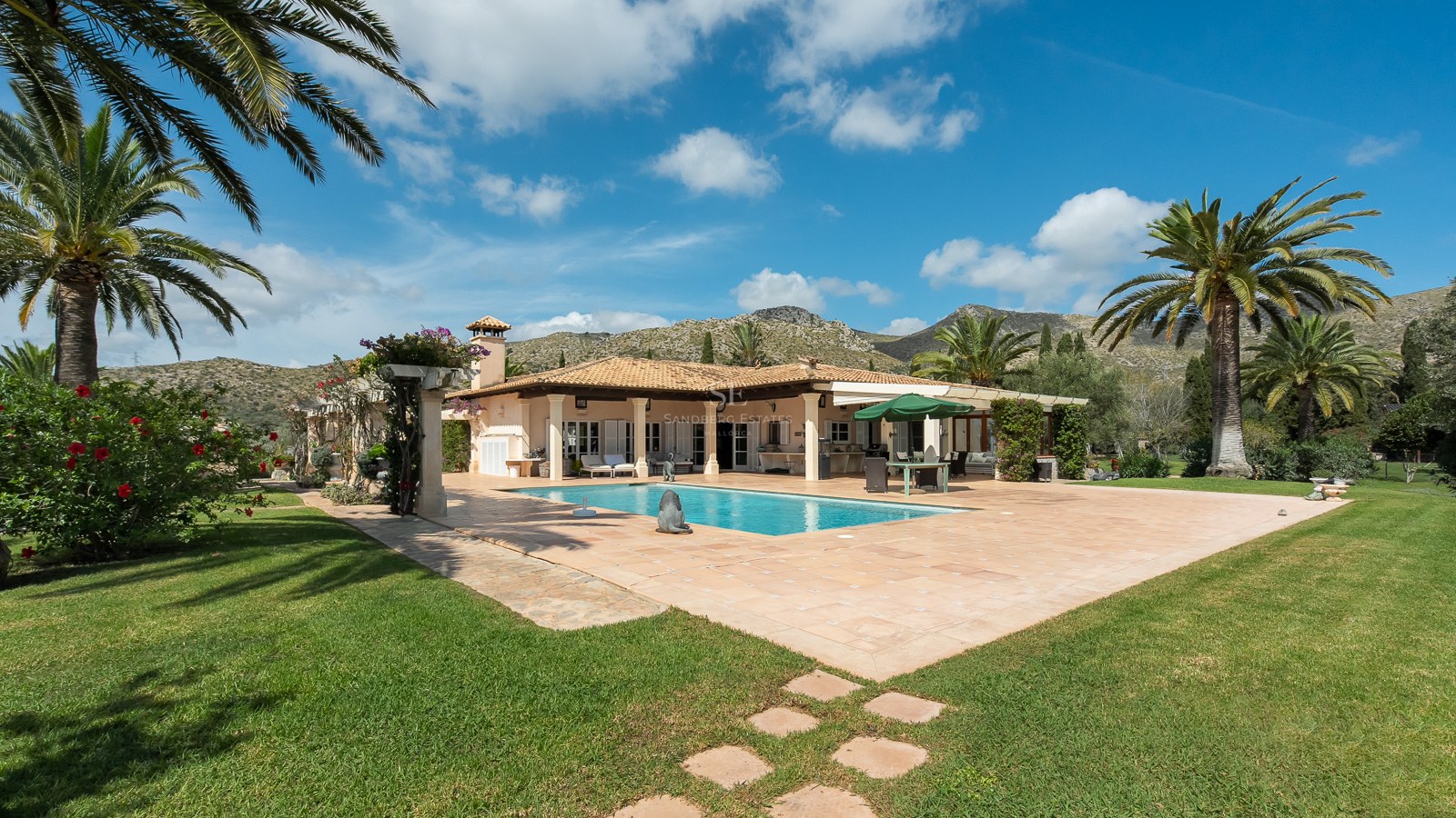 Spanische Villa mit rechteckigem Pool, Terrakotta-Terrasse, Palmen und Bergen im Hintergrund unter blauem Himmel.