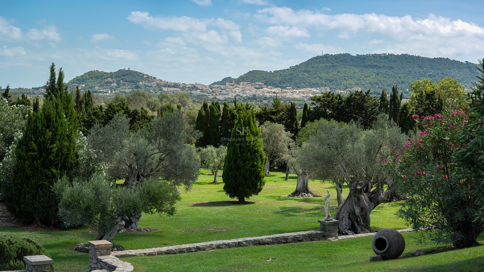 Üppiger mediterraner Garten mit alten Olivenbäumen und Steinmauern, Blick auf ein Bergdorf und Berge.