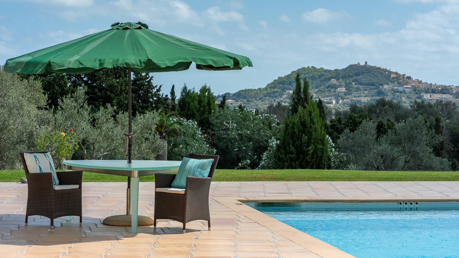 Terrasse au bord de la piscine avec chaises en osier sous un parasol vert, surplombant des jardins et des montagnes.