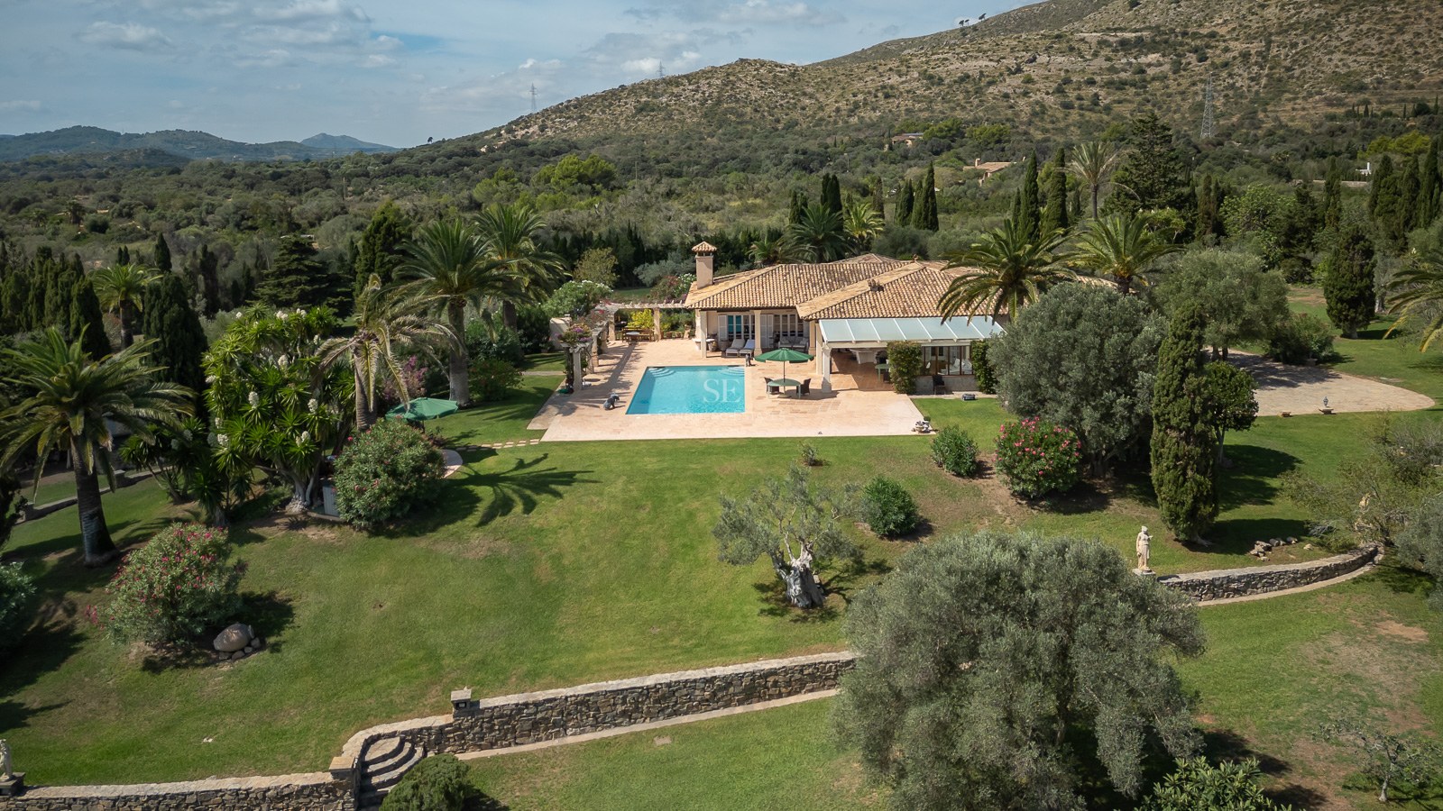 Vue aérienne d'une villa avec toit en terre cuite, piscine, grand jardin et montagnes en arrière-plan.