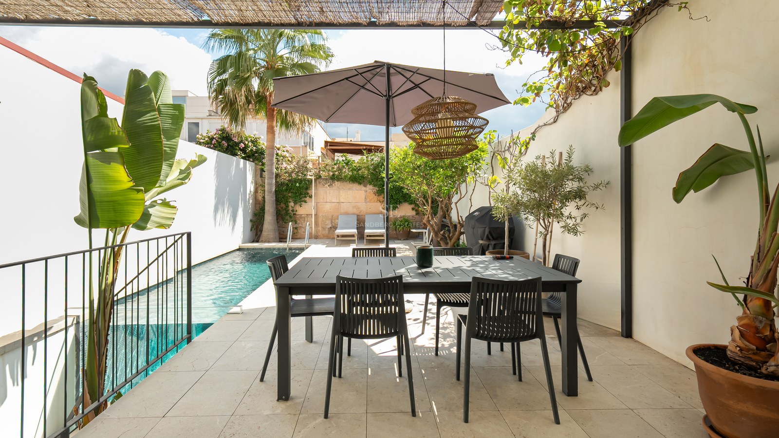 Terrasse moderne avec table à manger noire sous un parasol, à côté d'une piscine turquoise et de palmiers.