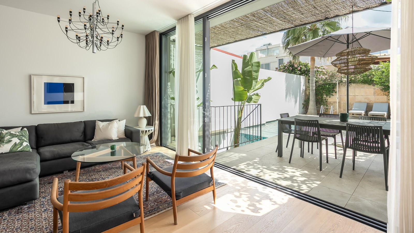 Salon lumineux avec canapé sombre et chaises en bois ouvrant sur une terrasse ensoleillée avec piscine et coin repas.