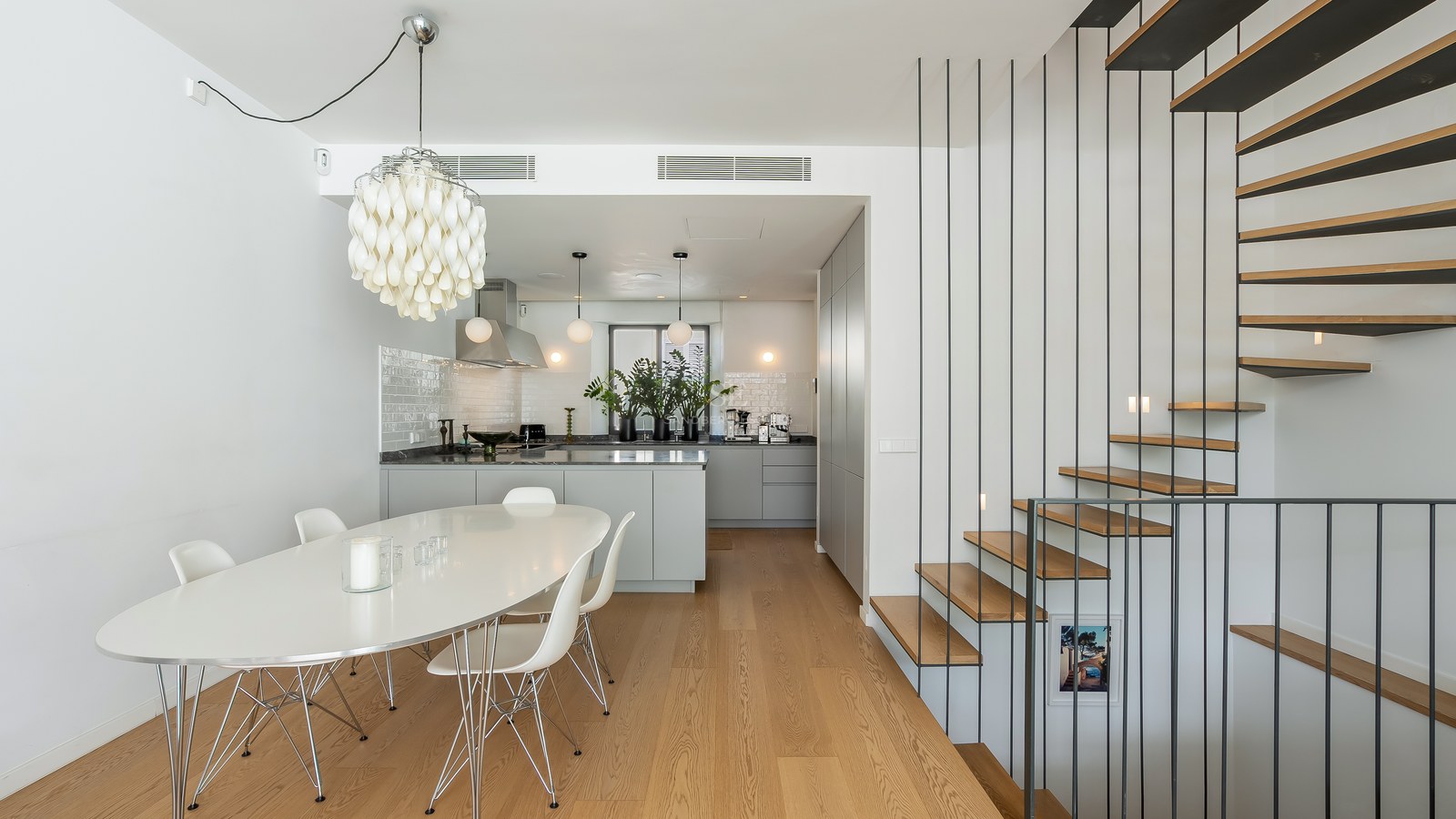 Salle à manger blanche avec table ovale, chaises Eames, lustre design, cuisine grise et escalier en bois et métal.