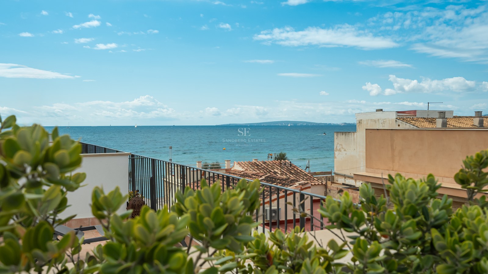 Vue sur la mer Méditerranée bleue depuis une terrasse avec une balustrade en métal noir et des toits en terre cuite.